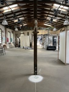 Die Installation ist in etwa 2 Metern Höhe  an einem Holzbalken der Halle befestigt. Die Skulptur hält  ein freischwingendes Lot, das direkt über einem Urglas angebracht ist. Im Urglas sind Wassertropfen. Daneben steht ein Wasserglas mit Pipette. 