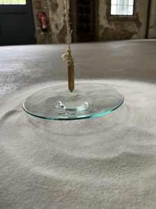 Ein Urglas am Boden mit einigen Tropfen Wasser darin. Es steht auf einer Fläche, die mit feinem, weißen Sand bestreut ist.